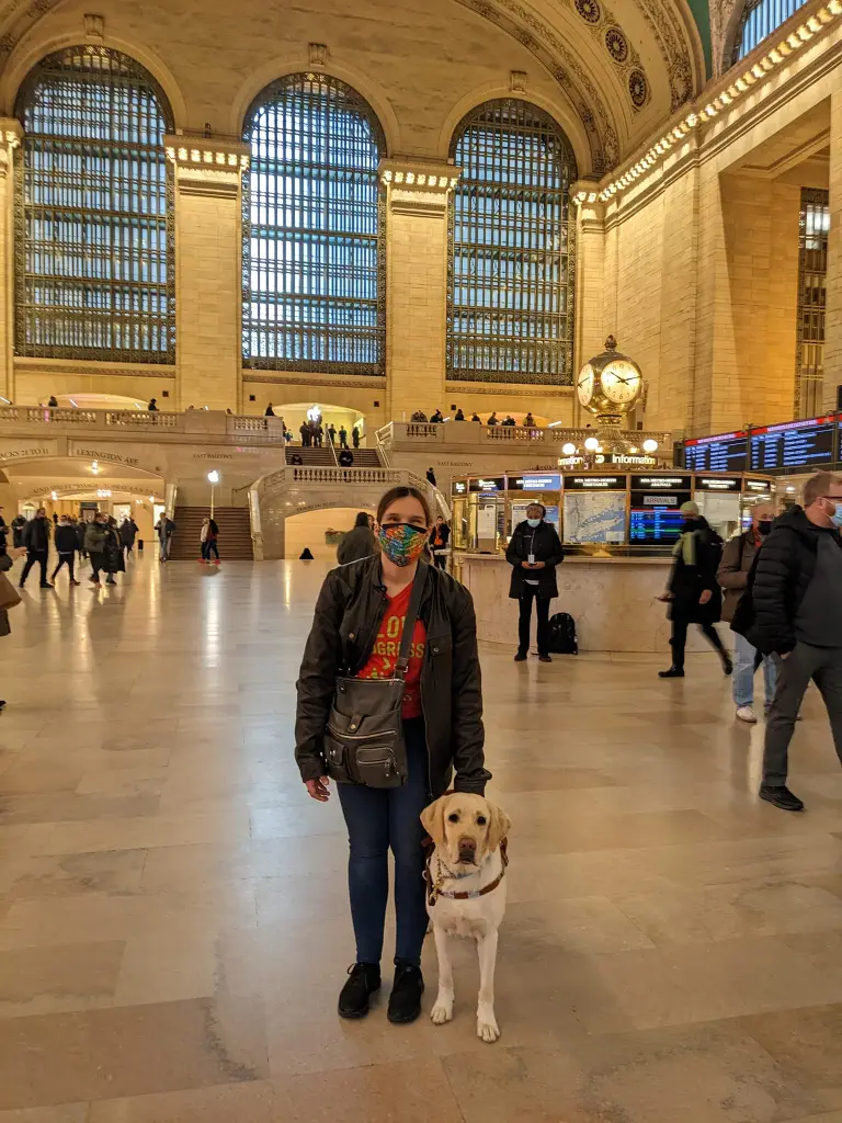 A masked woman in a red shirt is standing beside a yellow lab guide dog. Grand Central station is recognizable and visible in the background